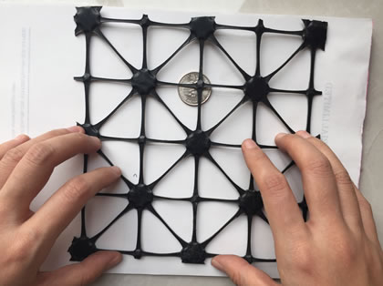 Two hands are pressing a piece of black triaxial geogrid.